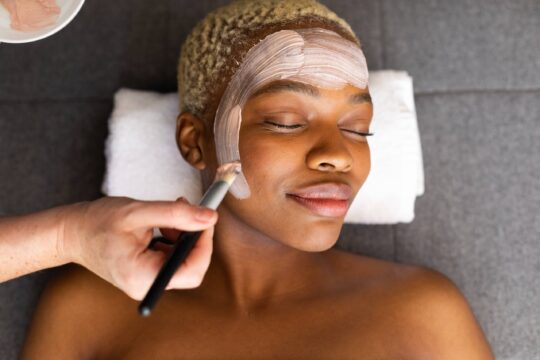 A woman receives a facial, with a person applying skincare product to the woman’s face using a brush